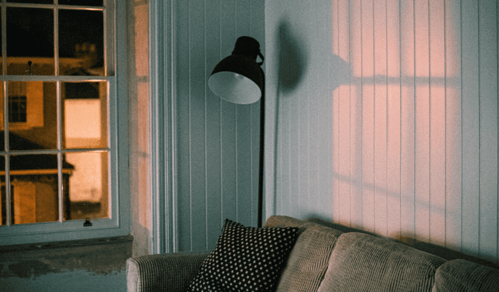 Sala aconchegante com sofá bege, almofadas decorativas e luminária de piso preta ao lado da janela refletindo luz suave na parede.