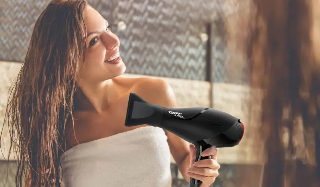 Mulher jovem e sorridente usando o novo secador Taiff profissional preto em frente a um espelho. Ela está segurando o secador na mão, secando seu cabelo molhado e enrolada em uma toalha, demonstrando praticidade e resultados rápidos.
